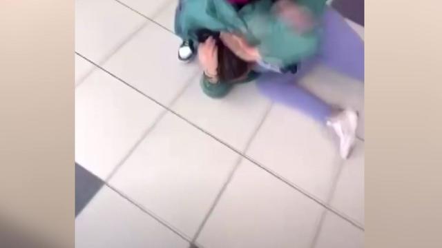 A teenage girl lays on the floor of a high school hallway, covering her head with her hands, as someone standing above her hits her. 
