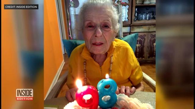 A photo of Alayne Skylar's mother posting in front of a birthday cake with lit candles in the shape of a 9 and an 8. 