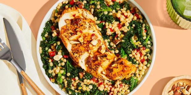 Plated garlic honey chicken atop pearl couscous with kale, roasted red peppers, and almonds, next to knife and fork and small red bowl with crushed almonds, all sitting on a green tablecloth 