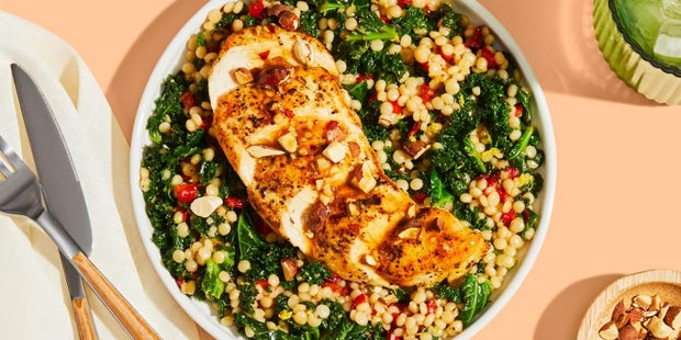 Plated garlic honey chicken atop pearl couscous with kale, roasted red peppers, and almonds, next to knife and fork and small red bowl with crushed almonds, all sitting on a green tablecloth