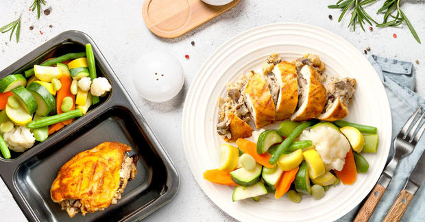 Plated Stuffed Chicken with Steamed Vegetables, off-center, next to black carryout container with identical meal separated into two sections 