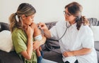 A woman holds a baby while a female healthcare worker presses a stethoscope to the baby's back 
