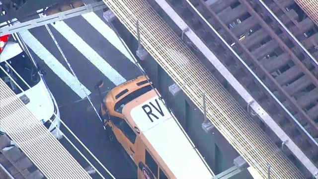 An aerial view of a school bus and an NYPD vehicle parked under elevated subway tracks. 