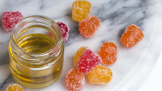 CBD gummy candy gumdrops in red, yellow, and orange surrounding a jar of hemp oil on a gray and white marble surface. High angle view. 