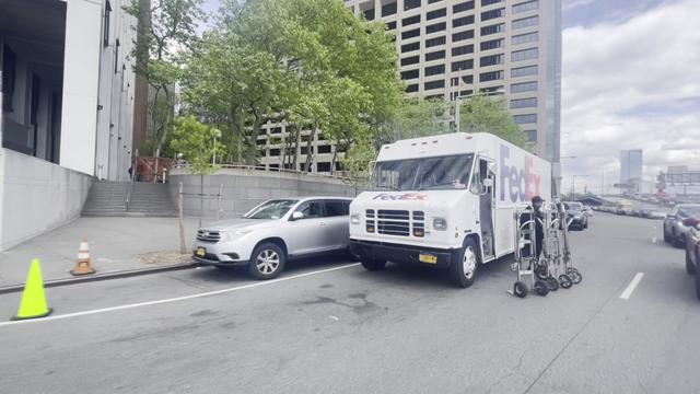 A FedEx truck sits double-parked on a street, partially blocking a lane, as the driver stands next to the vehicle with dollies. 