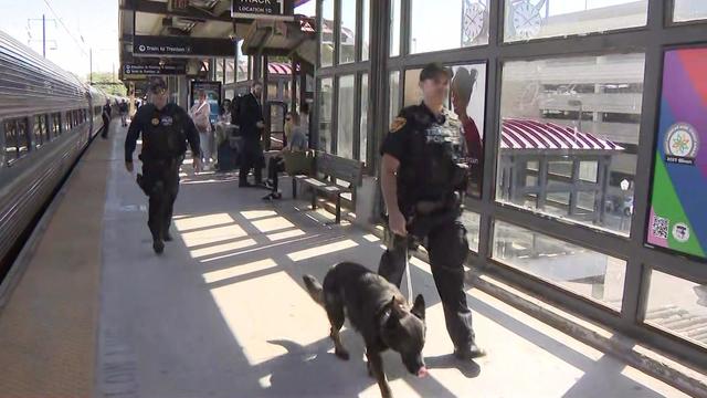 Two transit police officers, one walking a dog, walk along a train station platform. 