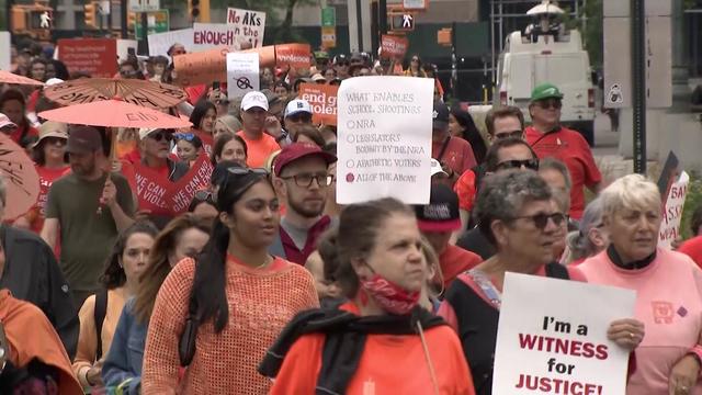 Dozens of people, most wearing orange, walk down New York City streets, some holding signs. 