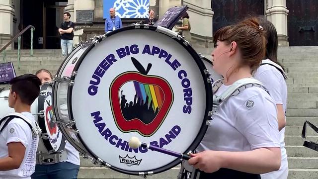 Bass drum players from the Queer Big Apple Corps marching band perform outside a cathedral. 