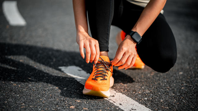 Closeup of woman knelt down on asphalt, tying laces on orange running shoes 