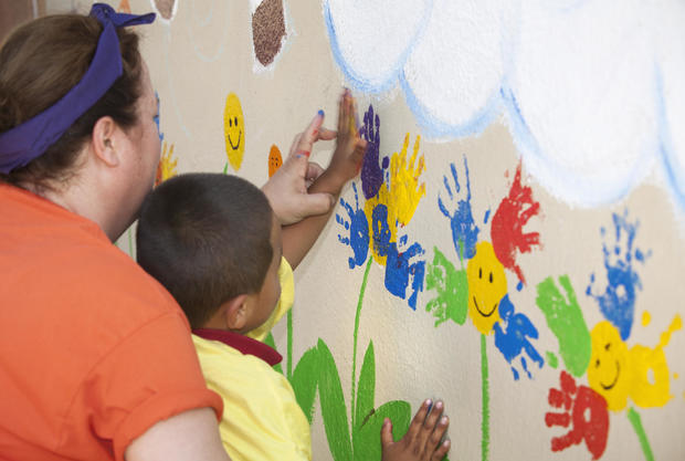 Kindergarten teacher helps a young boy paint a colorful mural using his handprint. 