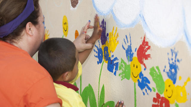 Kindergarten teacher helps a young boy paint a colorful mural using his handprint. 