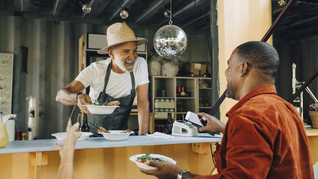 Smiling owner serving food while male customer doing contactless payment through smart phone 
