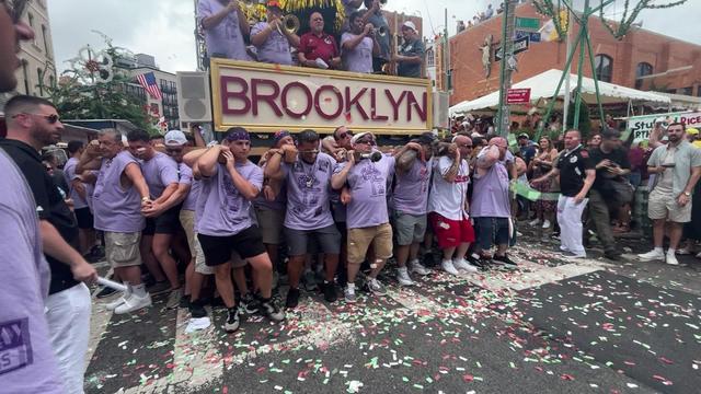 Dozens of men, most wearing light purple TV shirts, hold up a Giglio with a sign that says "Brooklyn." There is a band performing on the Giglio. Spectators line the streets. 