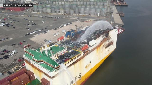 An aerial view of two boats spraying water on a cargo ship transporting dozens of vehicles. 