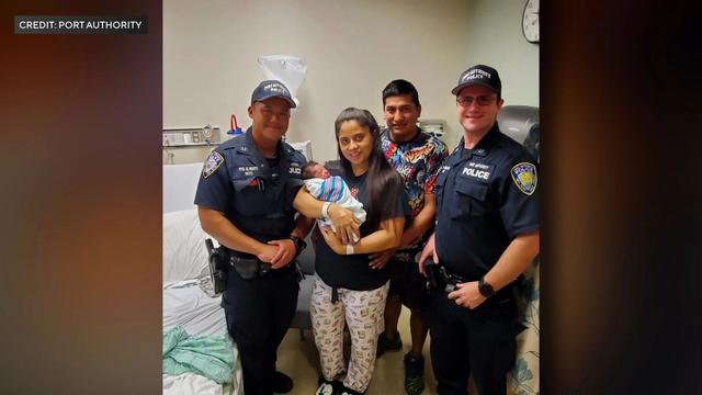 A mother holds a newborn baby in her arms in a hospital room. Her husband stands behind her and two Port Authority Police officers stand on either side of them. 