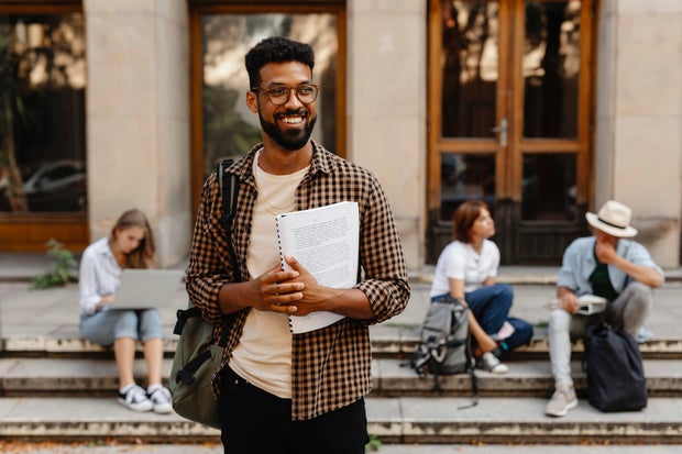 Happy young university student with book standing outdoors in front of campus. Back to university concept.