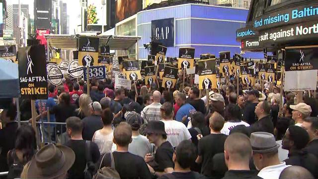 Dozens of people fill Times Square in front of a stage, many holding signs that say "SAG AFTRA ON STRIKE." 