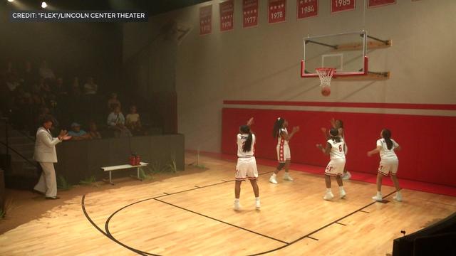 On a stage adapted to look like a basketball court, five young women in basketball uniforms play basketball. 