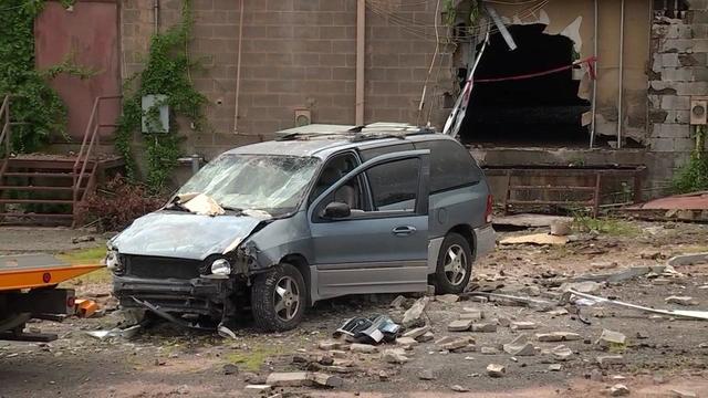 A mini van with a missing front bumper and hood and windshield damage sits in front of a vacant building with a large hole in the exterior wall. 