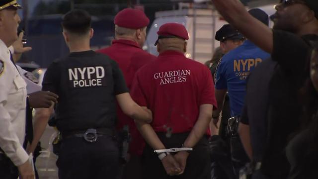 An NYPD officer leads away an individual, whose hands are zip-tied behind their back, in a Guardian Angels shirt and red beret. 