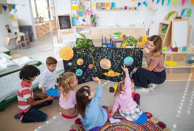 High angle view of pre school teacher showing poster with planets to children indoors in nursery, Montessori education. 