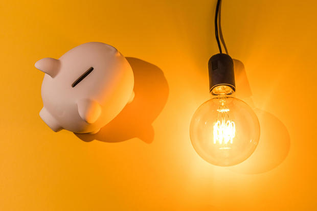 Piggy bank next to lit light bulb on pink background. Concept of electricity price, crisis, money, saving and power energy. 