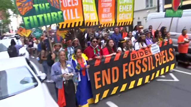 Dozens of people at the front of a protest carry a large banner reading "Biden End Fossil Fuels." 