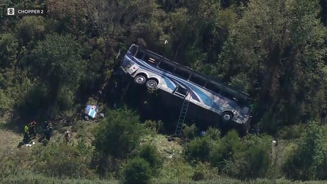 An aerial shot of a charter bus laying on its side in a wooded area on the side of an interstate. 