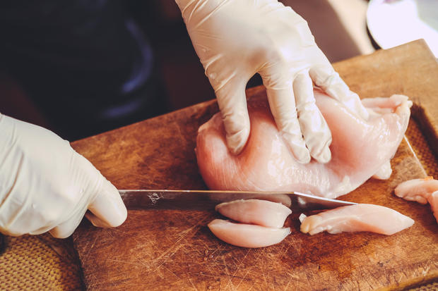 Hands in gloves slicing raw chicken fillet. 