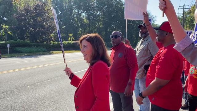 Gov. Kathy Hochul holds a strike sign and stands with picketing UAW members on the side of a street. 