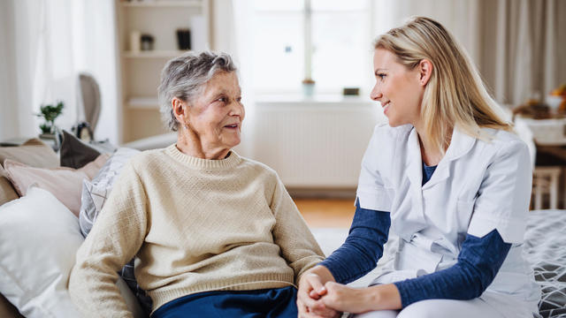 A health visitor talking to a sick senior woman sitting on bed at home. 