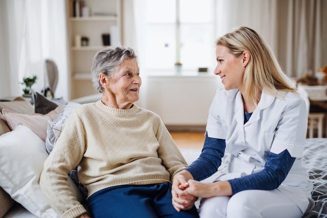 A health visitor talking to a sick senior woman sitting on bed at home. 