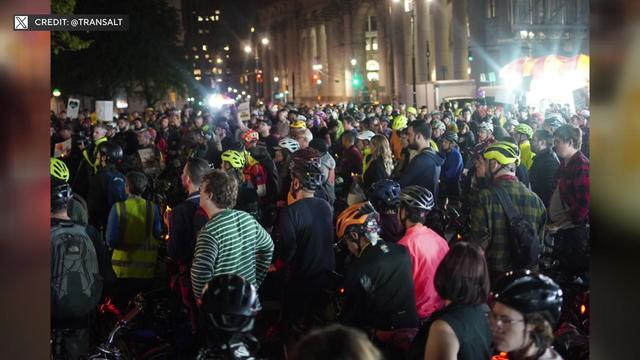 Hundreds of people, many wearing bike helmets, fill a New York City street. 