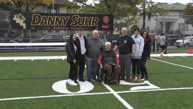 Firefighter Danny Suhr's loved ones stand on a football field in front of a sign that says "Danny Suhr Memorial Field." 