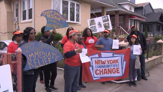 About a dozen individuals stand in front of homes in Queens holding a "New York communities for change" banner and signs reading, "Sandy survivors deserve community engagement." 