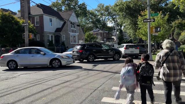 Multiple vehicles wait in an intersection and in a crosswalk as children wearing backpacks and an adult cross the street in another crosswalk. 