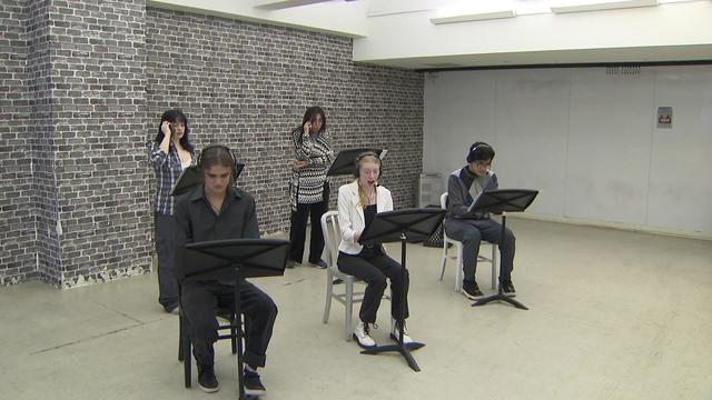 Three students sit on chairs in front of music stands and two students stand in front of music stands behind them while rehearsing a play. 