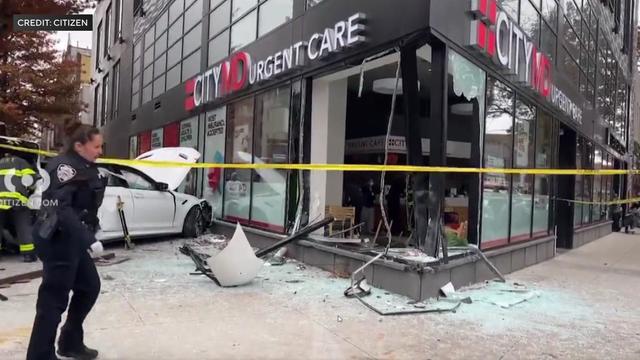 Crime scene tape blocks off the sidewalk outside a CityMD. A large window has been broken and shattered glass covers the sidewalk. Nearby, a white sedan sits on a sidewalk up against the building with its hood up. 