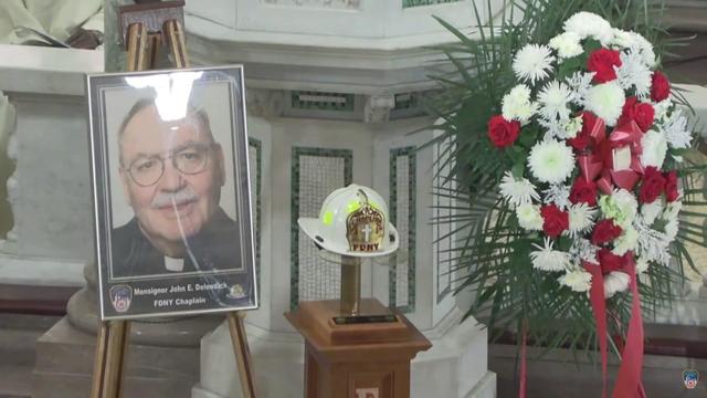 An enlarged photo of FDNY Chaplain John Delendick is displayed on an easel at his funeral, alongside his FDNY chaplain helmet and a wreath of flowers. 