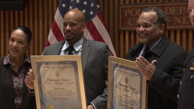 Retired NYPD officers Axel and Kenny Dodson hold framed award certificates and pose for photos with NYPD officials. 