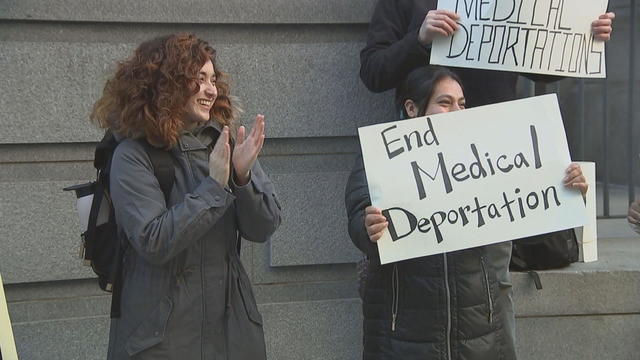 A woman claps after the vote, another holds a sign that says End Medical Deportation 
