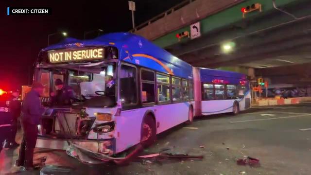 An MTA bus with extreme front-end damage sits in an intersection while investigators examine it. 