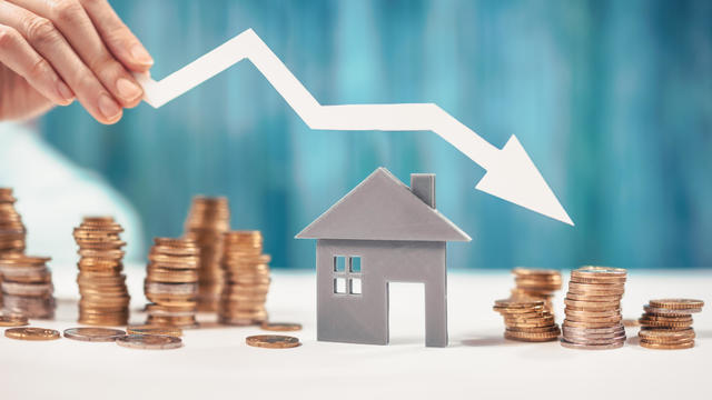 Woman holds an arrow down over model of the house and stack of coins. 