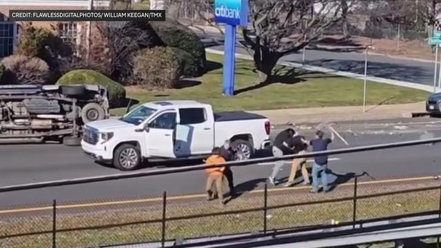 A white pickup truck with the driver's side door open sits in the middle of a street near another vehicle that has been tipped on its side. On the edge of the street, five individuals appear to be involved in an altercation, including one individual wield 