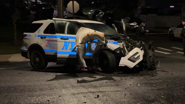 An NYPD patrol vehicle in an intersection with severe front-end damage and fire damage. 