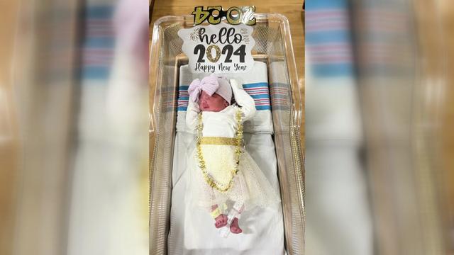 A newborn baby dressed in a sparkly white-and-gold dress lays in a hospital bassinet adorned with a "hello 2024 happy new year" sign. 