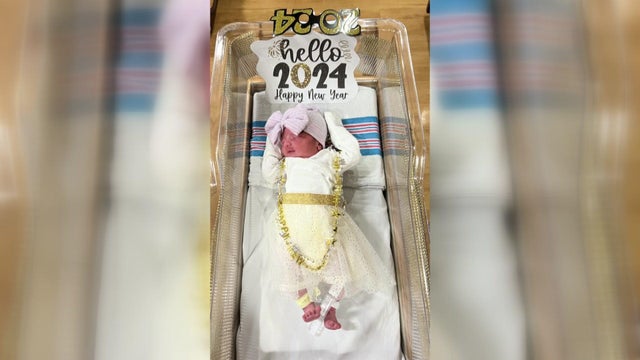 A newborn baby dressed in a sparkly white-and-gold dress lays in a hospital bassinet adorned with a "hello 2024 happy new year" sign.