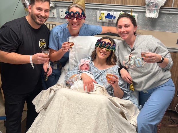 Two medical staff members pose with parents and a newborn baby in a hospital bed. One medical staff member and the mother wear novelty 2024 glasses and all four hold cups of what appears to be sparkling cider.