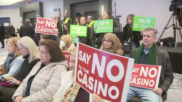 Individuals sit and stand inside a ballroom during a meeting. Some hold red signs reading, "Say no to the casino," while others hold green signs reading, "Yes to Sands." 