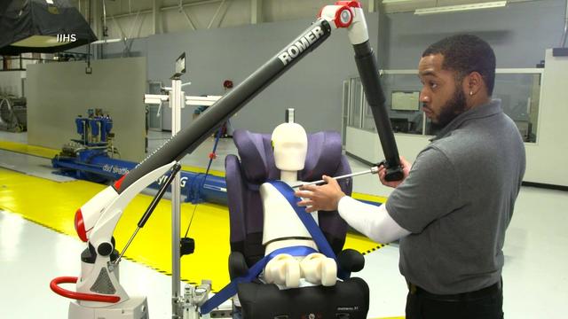 A man tests a booster seat with a child-sized crash test dummy strapped into it. 